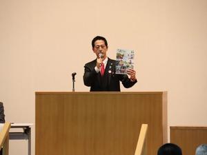 女性と農業が表紙となった広報よしかわを手に壇上から挨拶をする市長
