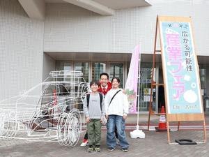 産業フェアの看板とフレームカーをバックに来場した子供達と記念撮影する市長