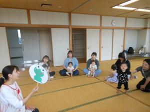 保育士が緑の風船の絵を持って歌っているのを一緒に見ている様子