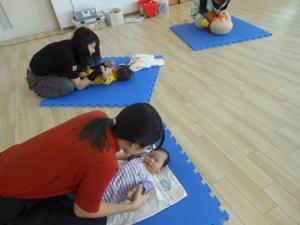 二組の親子がそれぞれ寝転がっている自分の子どもに顔を近づけて見つめているところ